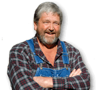 fred schouten met een grijze baard en kort grijs haar draagt een geruite overhemd en blauwe denim overalls, terwijl hij glimlacht en zijn armen over elkaar houdt.
